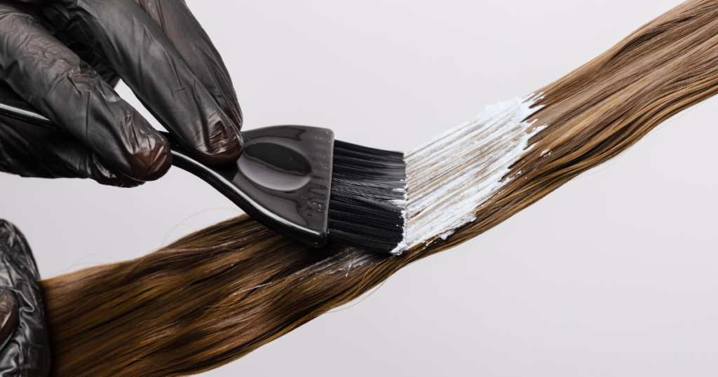 A gloved hairstylist applying white hair dye to a brown hair strand with a tinting brush