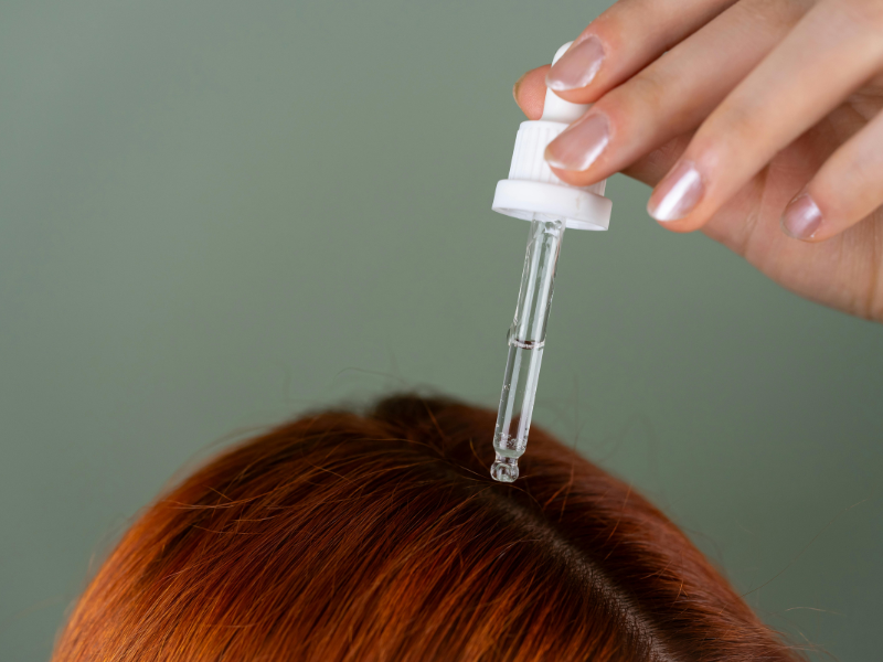 A woman applying clear serum drops from a dropper to her scalp to remove hair dye from skin