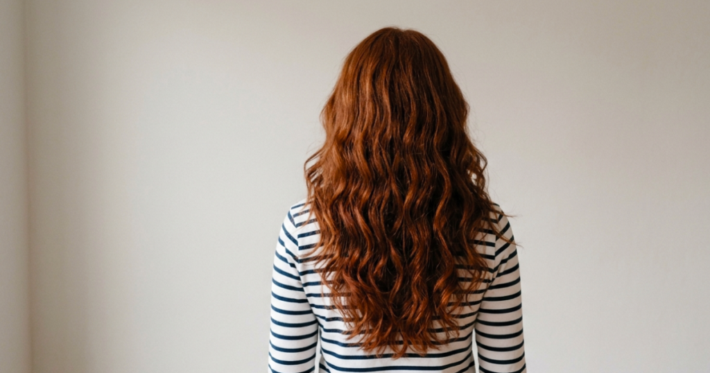 Woman with long, voluminous dark copper red wavy hair shown from behind, wearing a striped top