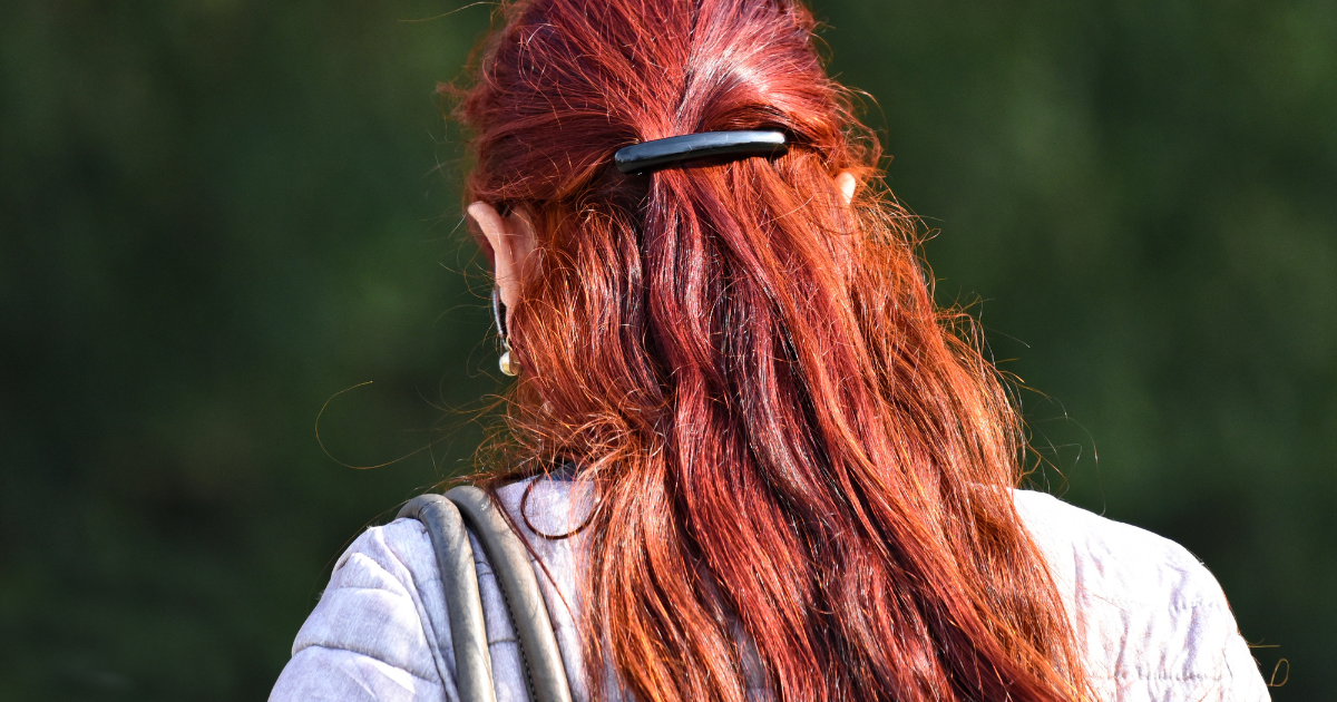 Woman with vibrant red hair secured with a black barrette, seen from behind against a green outdoor background