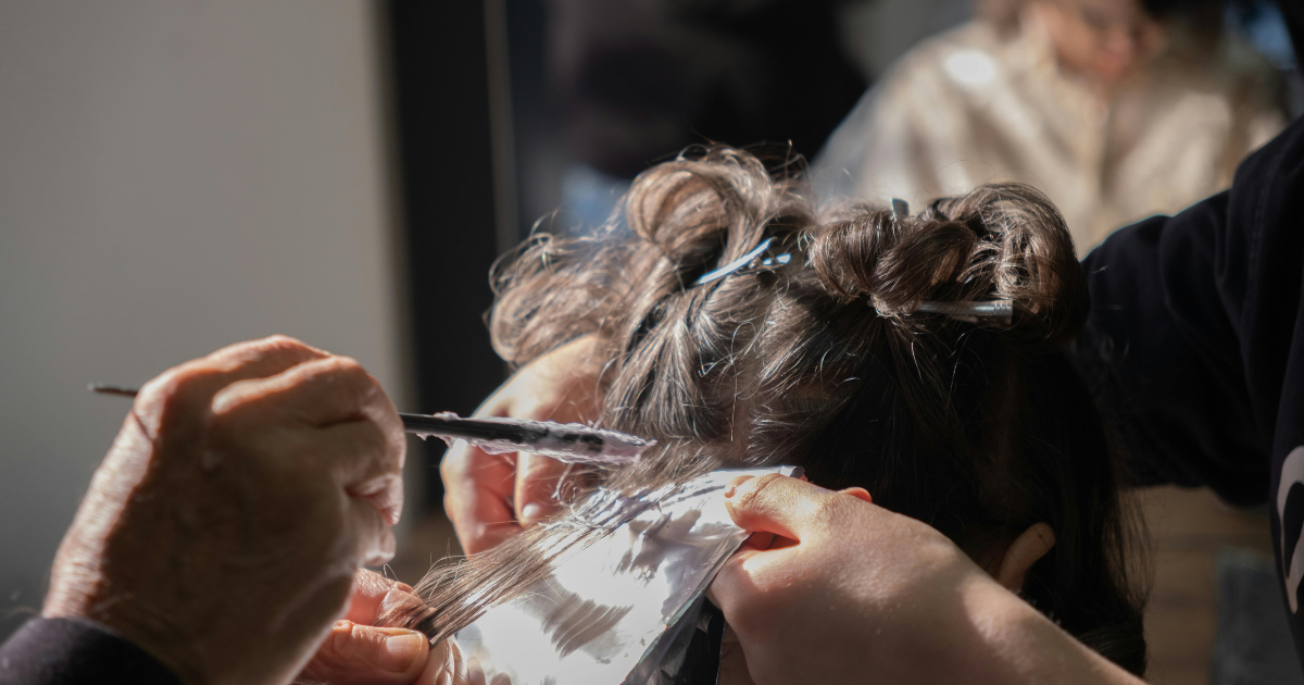 Close-up of a stylist painting hair color onto foil-wrapped sections of dark brown hair, demonstrating a professional coloring technique