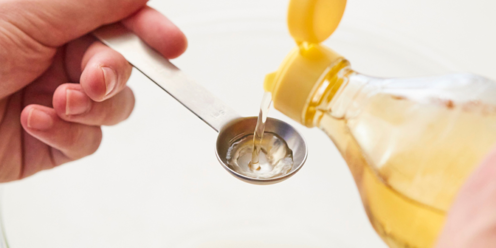 Pouring a measured teaspoon of oil from a bottle, ready to use as a gentle home remedy for removing black hair dye from skin