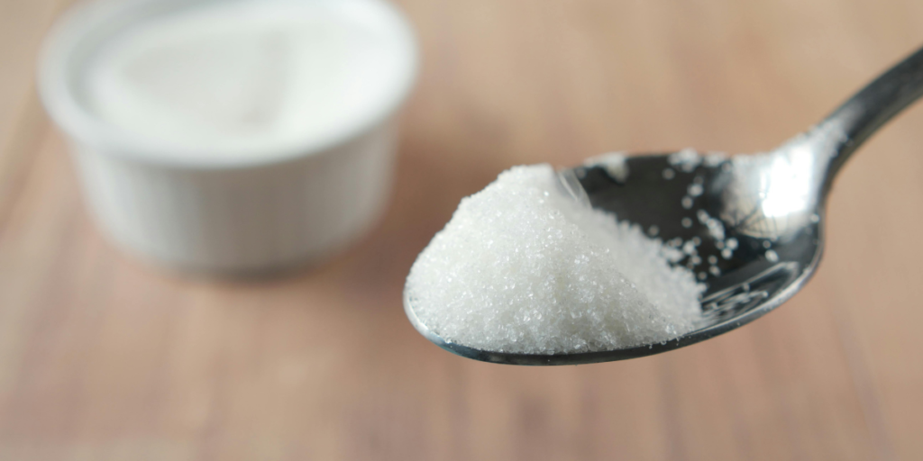 A heaped spoonful of white granulated sugar with a small bowl of sugar in the background, key ingredients for a DIY scrub to remove black hair dye from hands