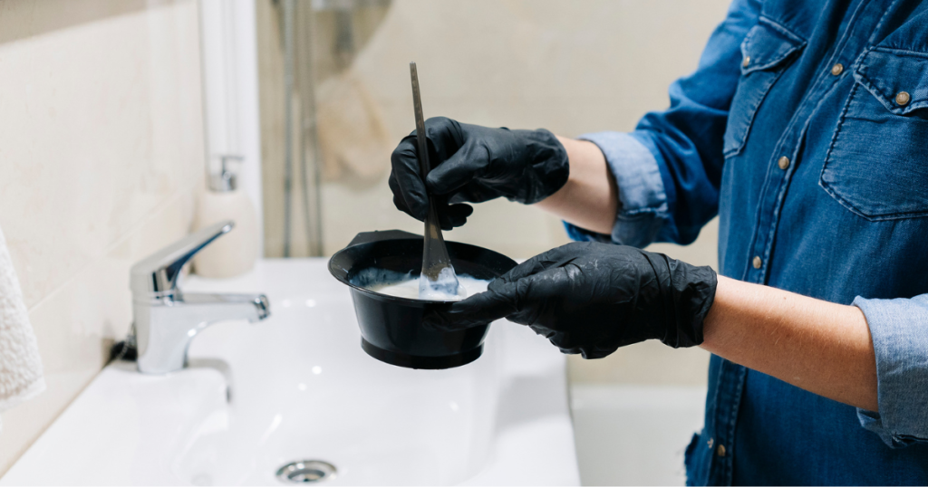 A person wearing black gloves mixing black hair dye in a bowl over a white bathroom sink, preparing for an at-home hair coloring session.