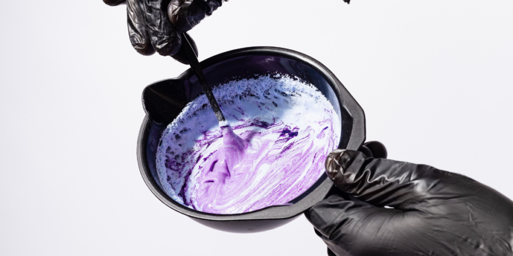 Hands in black gloves stirring freshly mixed purple hair dye in a black mixing bowl, showing the creamy consistency ready for at-home application.